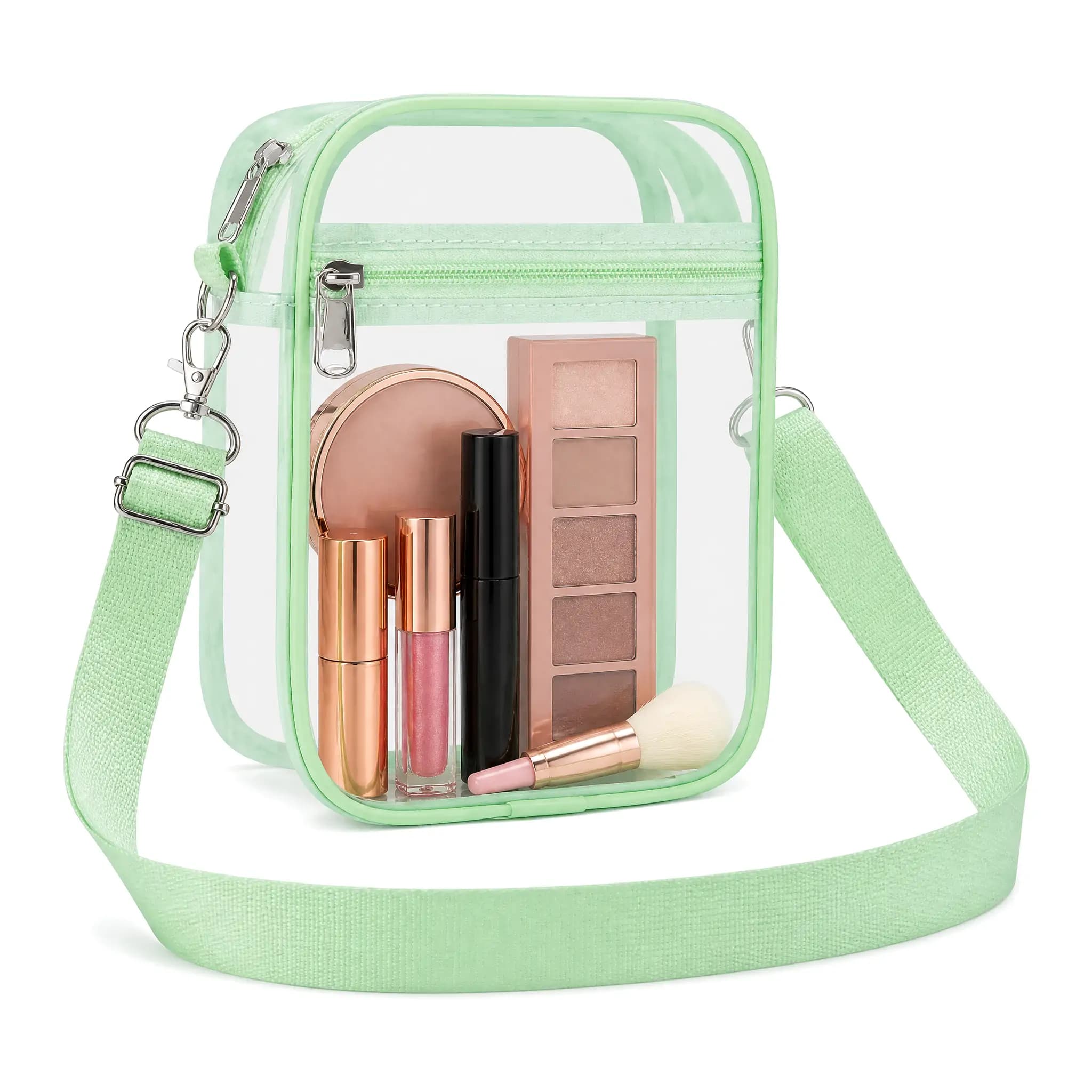 Clear PVC crossbody stadium bag 8.3 x 6.6 x 2.8 in with mint green binding, metal snap-hook strap and two zip compartments, shown filled with cosmetics on a white background