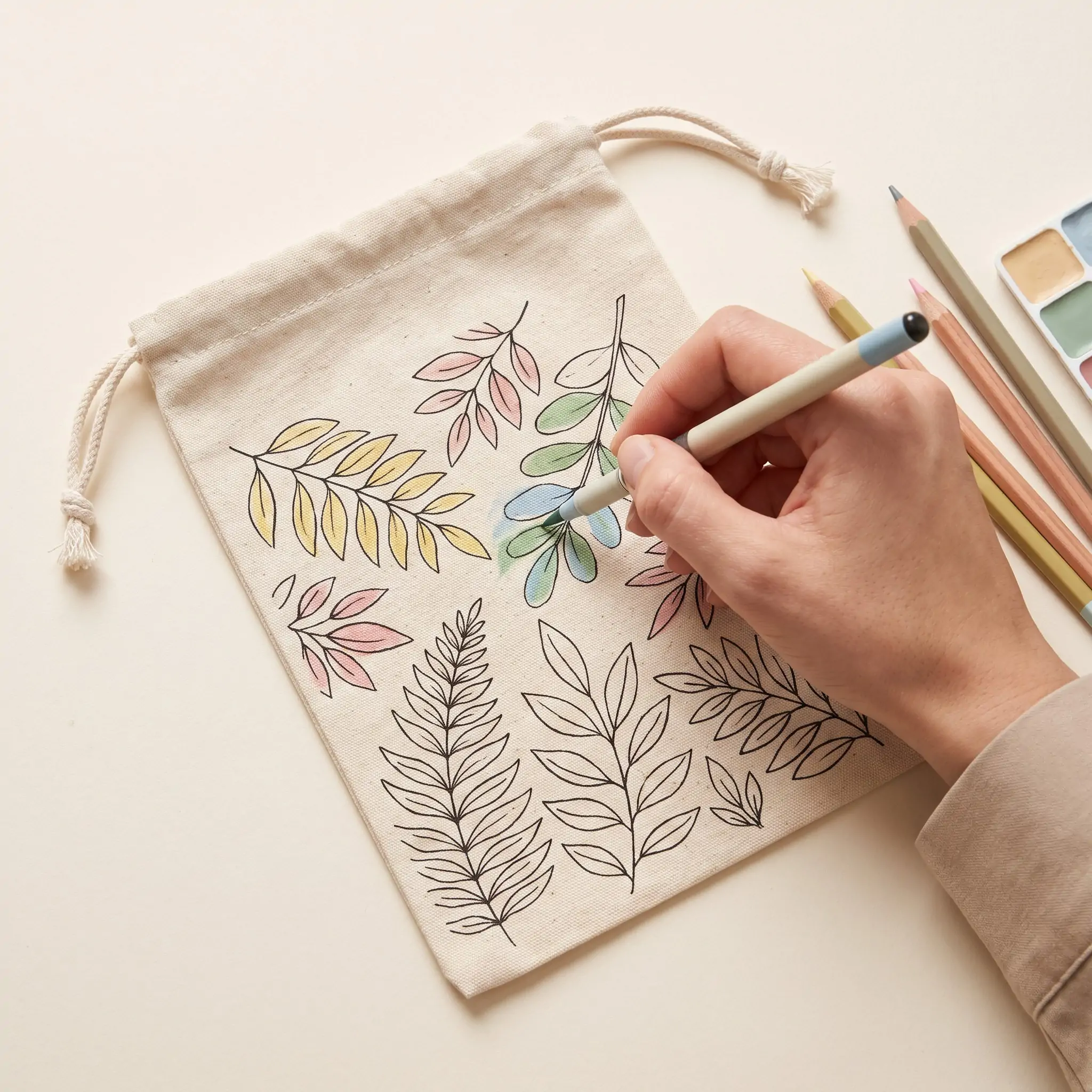 Natural canvas drawstring pouch 15x20cm with botanical line-art floral print, a hand coloring the artwork with a marker, and a knotted cinch cord